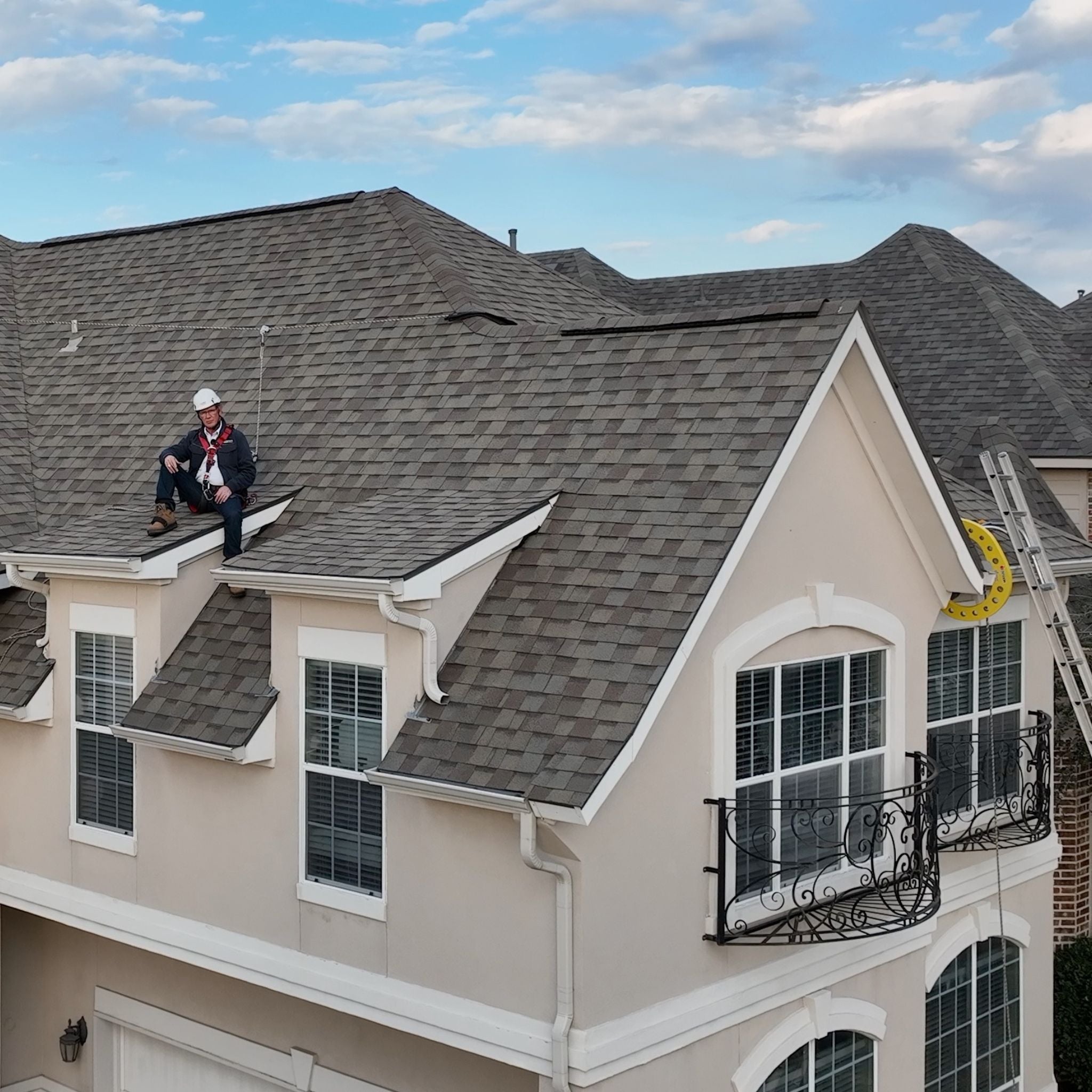 Roof worker sitting on ledge of residential sloped roof wearing white CAMP Safety helmet, roof safety harness PPE hooked into a non-penetrating roof anchor.