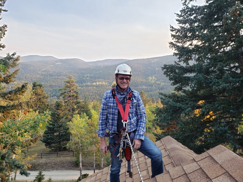 Roof worker at peak of sloped roof wearing roof helmet, PPE, and roof safety harness with mountains in background.