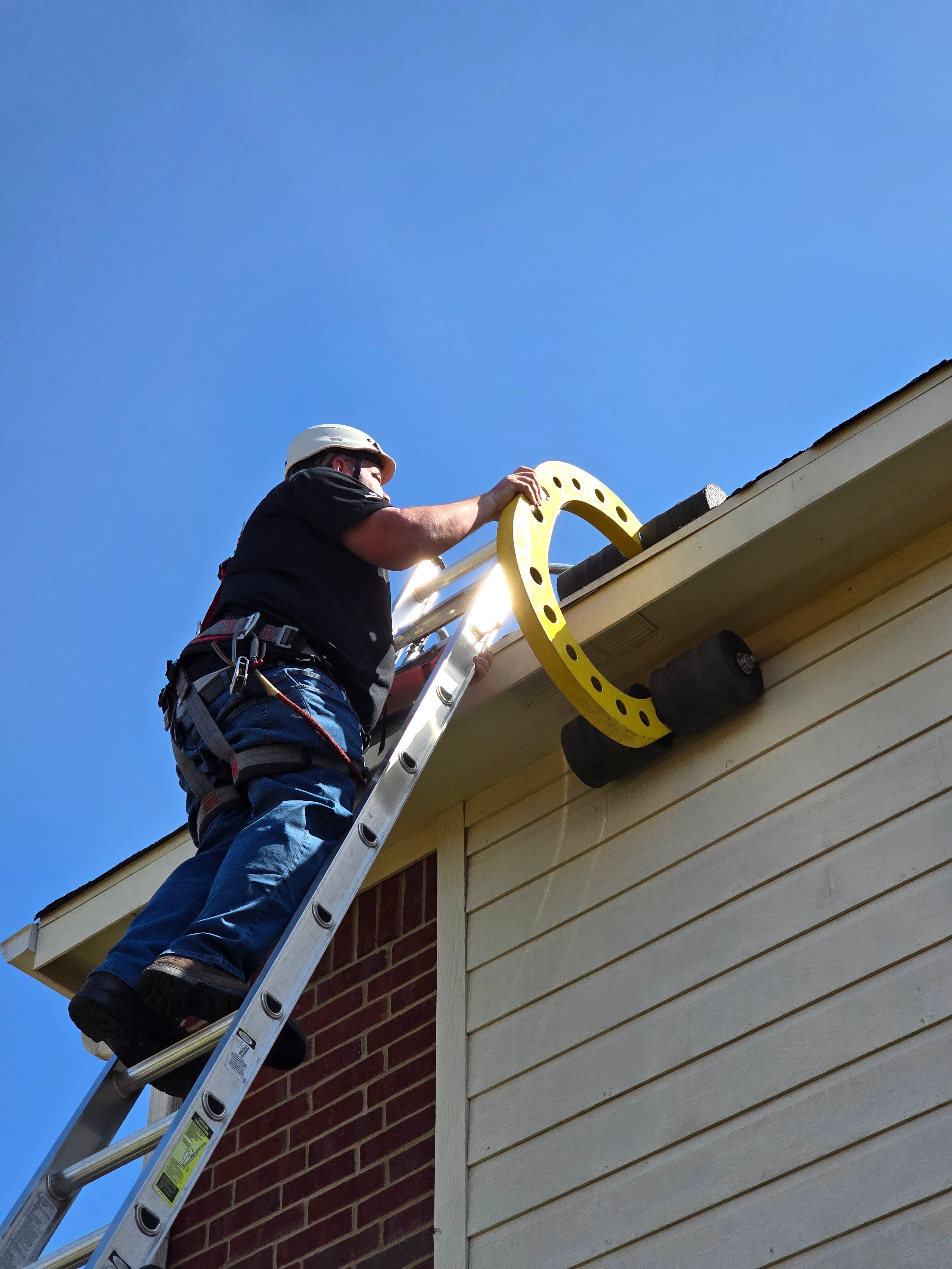 Roof worker at the top of a ladder holding an Eveook on the eave of a residential sloped roof home wearing roofing PPE, roof safety harness and roofing helmet.