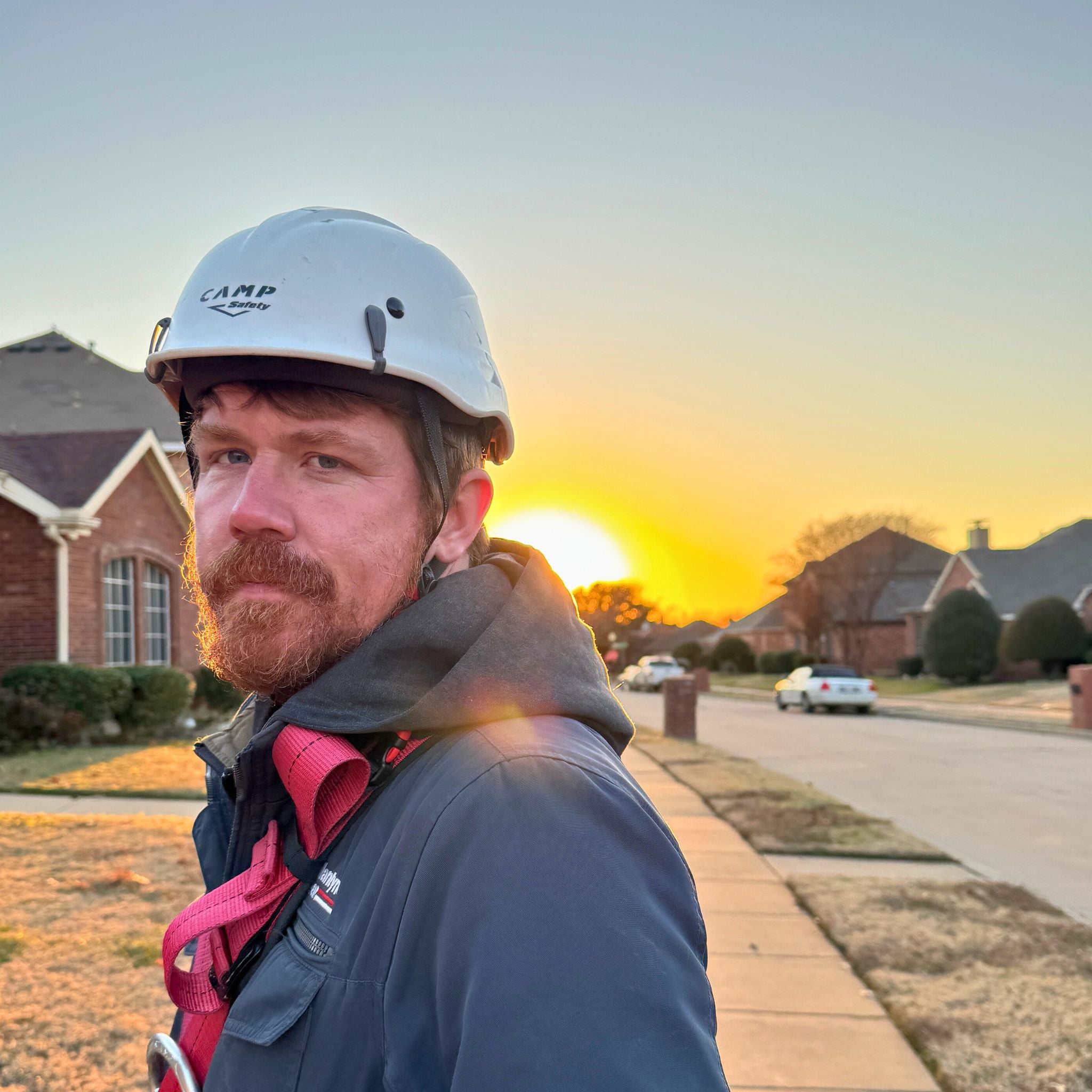 Roof worker wearing CAMP safety white helmet PPE and roof safety harness with sunset in the background.