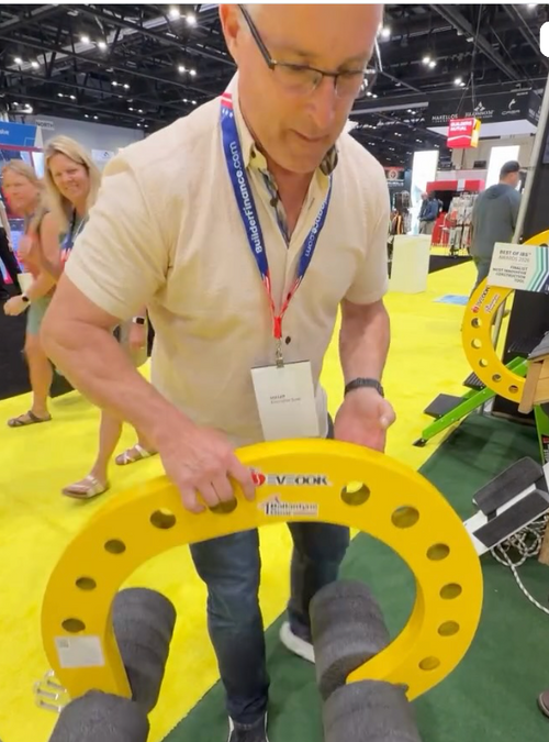 Man at a trade show holding a yellow tool, with a blurred background of other attendees and booths.