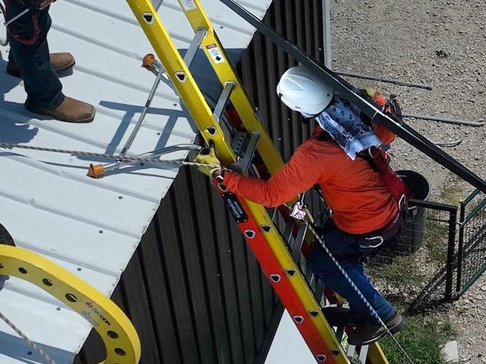 Solar worker climbing ladder carrying solar panel and using Eveook fall protection system on a standing seam metal sloped roof.